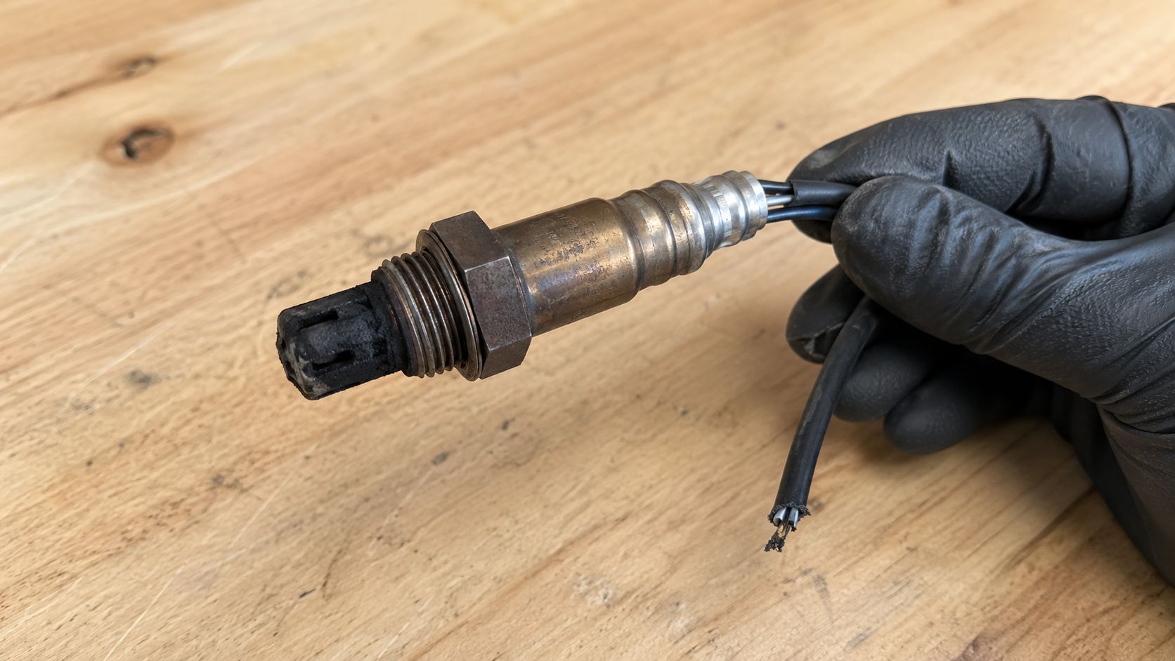 A hand in a black glove holds a used, carbon-fouled oxygen sensor with a cut electrical wire.